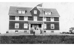 一张黑白照片，三层楼的房子，深色油漆和白色装饰。这所房子坐落在一大片土地上，房子前面有一个人站在旗杆旁。