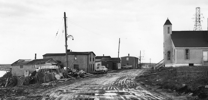 A black and white photo of a muddy road with wooden houses on one side and a white church on the other.