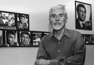 吉姆·克拉克(Jim Clarke)站在一面墙前的黑白照片，墙上挂着几张默罕默德·阿里(Muhammad Ali)的照片。