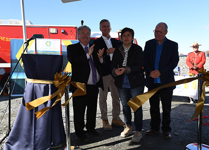 一张Lloyd Hines, Steven Dean, Maryse Belanger和Terrance paul的照片。他们站在一个大帐篷下的阴凉处。海恩斯在鼓掌，其他人开心地笑着。贝朗格刚刚剪下了闪闪发光的金色丝带。在背景中，你可以看到一辆应急响应车辆和一名身穿红色军服的皇家骑警，像一个服务员一样站在烈日下的桌子后面，专注地对着人群微笑。