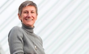 照片上，一位身穿灰色毛衣的微笑女子站在一座白色条纹建筑前。