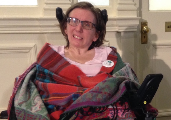 一个女人穿着粉红色的衬衫，戴着眼镜，盖着一条毯子，坐在轮椅上。