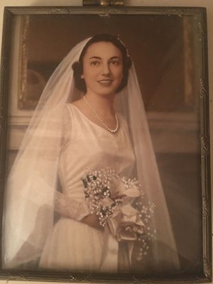 蒂姆美丽的妈妈在婚礼当天的照片。她穿着一件长袖白色连衣裙，戴着一串珍珠，戴着长面纱，手里拿着一束花。