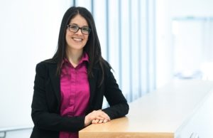 一个亮粉红色上衣和黑色毛衣的微笑的年轻女子靠在桌子上，靠在相机上。