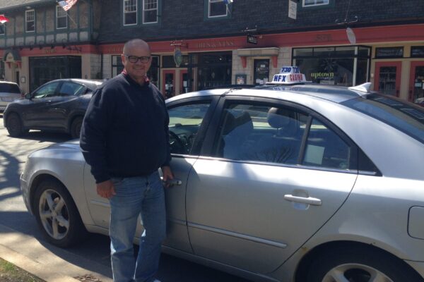 A smiling man stands next to a taxi on a sunny day. The car is a silver sedan with a taxi roof light. It's parked along Young Street in the Hydrstone.