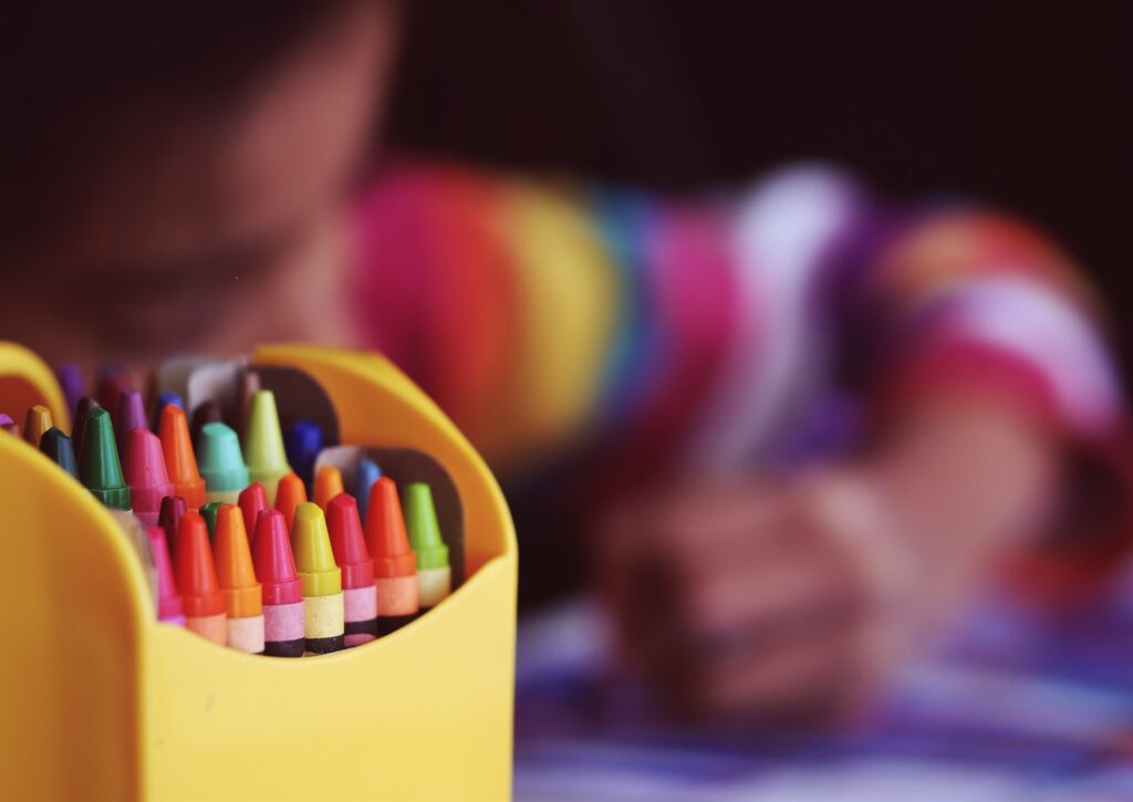 A box of coloured crayons stands in the foreground while a child is slightly out of focus in the background colouring on a piece of paper.