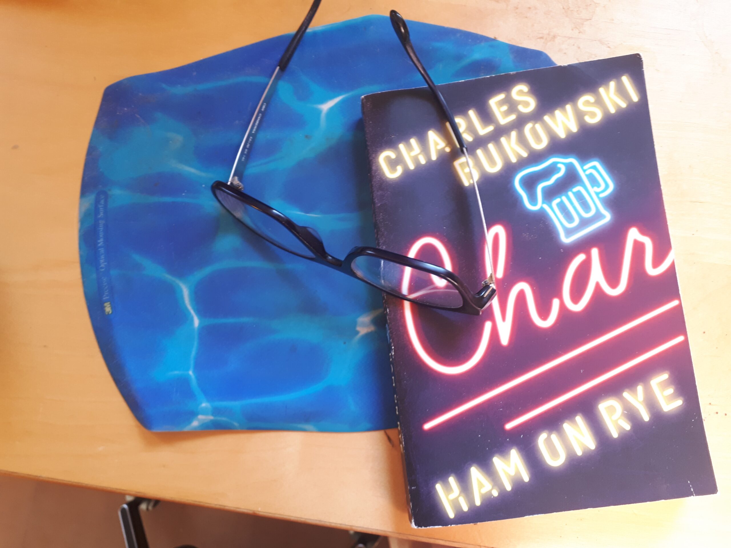 一本查尔斯·布可夫斯基的《黑麦火腿》放在鼠标垫上，上面放着一副眼镜。