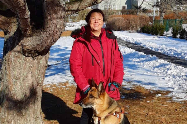 一位身穿红色冬衣的妇女站在一棵大树旁，带着她的狗——一只德国牧羊犬。在背景中，有一些雪覆盖着地面