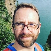 一个戴着眼镜，穿着亮色条纹衬衫，身后是大海的男人。