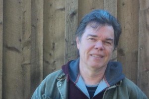 亚瑟·布尔(Arthur Bull)是一个黑发男子，对着镜头微笑，穿着一件绿色夹克，搭配深蓝色和酒红色连帽衫，站在一面木板墙前。