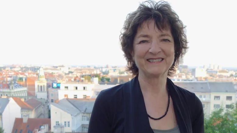 一个微笑的女人，戴着黑色西装外套和深色项链的短而深色的头发似乎站在屋顶上，在相机上微笑。