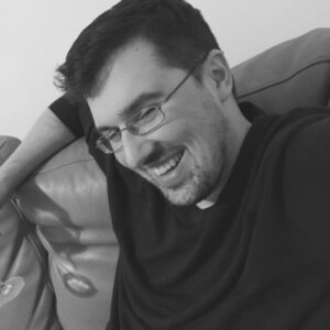 一张乔·卡伦坐在沙发上大笑的黑白照片。他笑容可掬，深色头发，戴一副金属框眼镜。就在镜头边缘，你可以看到一个可能是葡萄酒杯的底部。