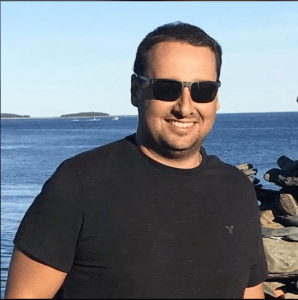 在这张以蓝色海洋为背景的Facebook照片中，警长迈克·萨克戴着黑色墨镜，穿着黑色t恤，对着镜头微笑。