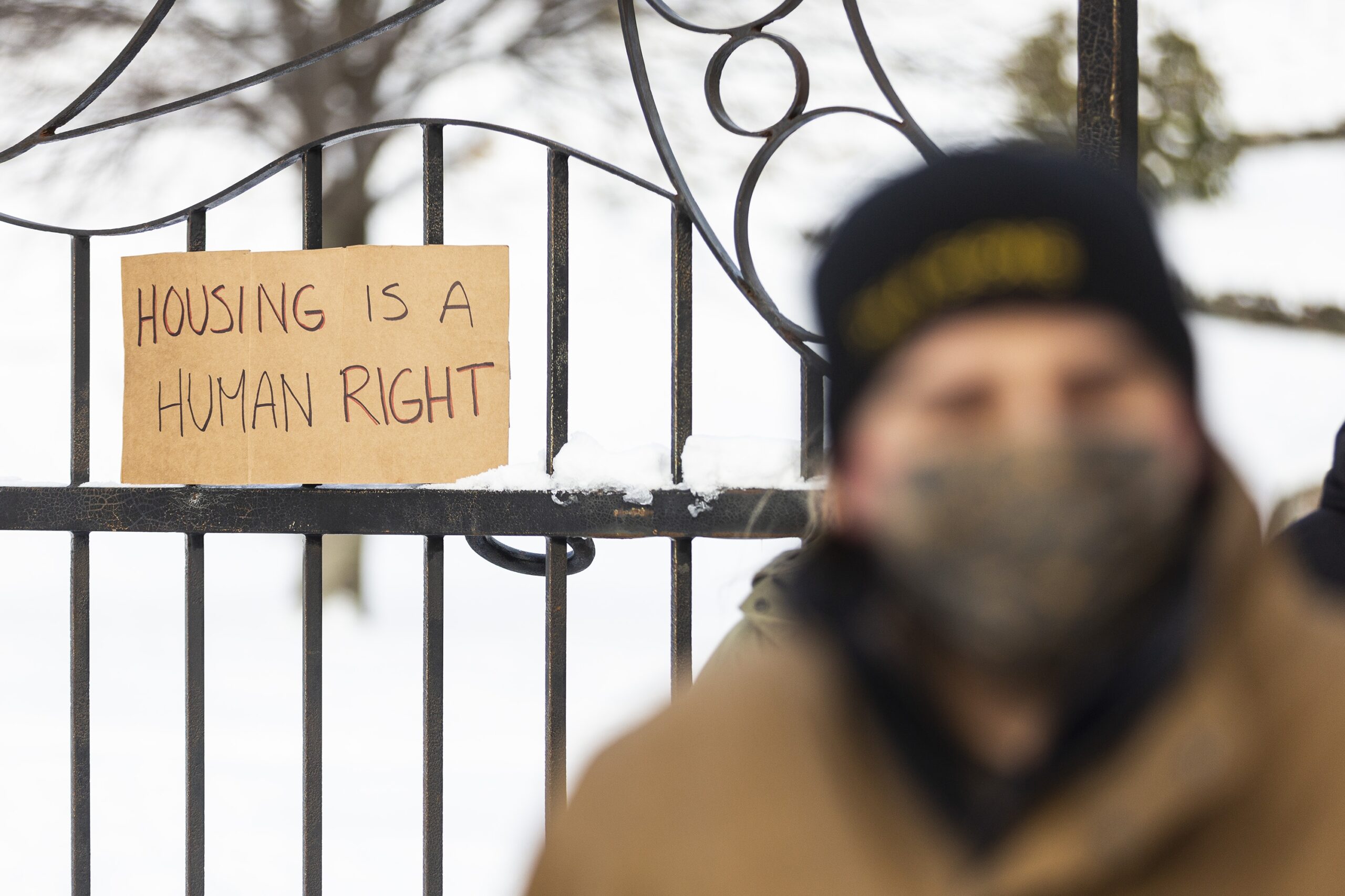纸板标志阅读住房是一个人权在冬天的锻铁围栏上被提出。在前景中，非常模糊，你可以看到一个穿着冬天的外套，拓和面料面膜的男人。