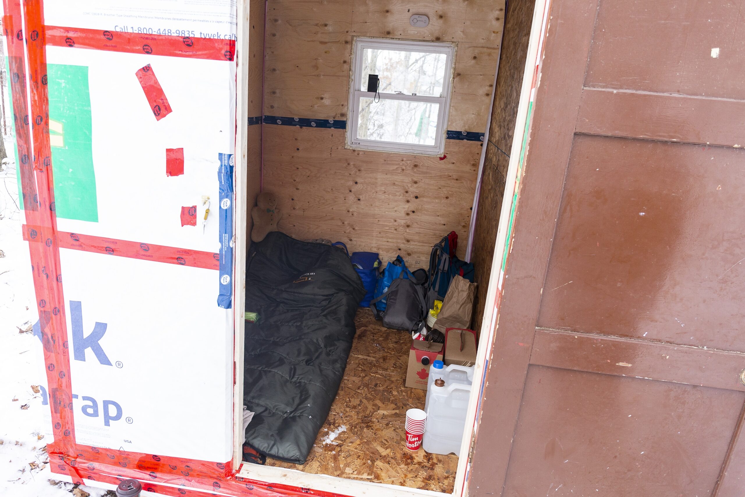 避难所内部的照片，这是一张双人床的大小。背面有一个木门和一个小窗户。里面是由刨花板和胶合板制成的，你可以看到一个睡袋和地板上的一些个人小便。