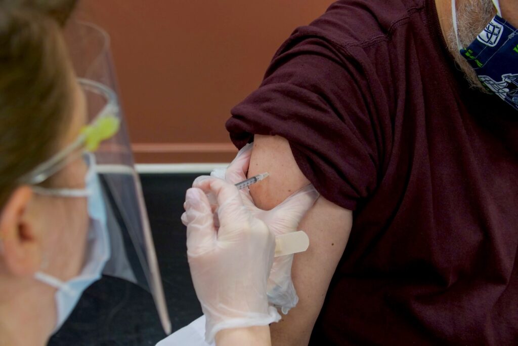 A nurse gives a vaccination to a man wearing a burgundy sweater with the sleeve rolled up