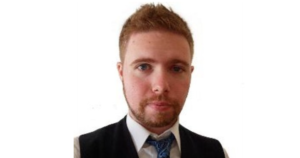 一个严肃的留着胡子的年轻人，穿着西装，打着领带，背景是白色的，他看着镜头。