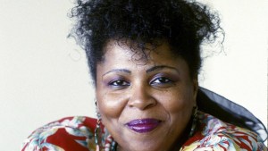 玛克辛泰恩。一个中年黑人妇女的特写脸。白色背景。她眼睛带着微笑，化着紫色的妆。她的头发是黑色的。她的肩顶露出了画框的底部，露出了花布