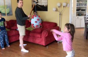 奥利维亚·希利登陆冠状病毒piñata的照片。她的爸爸杰夫拿着piñata。她的弟弟利亚姆站在爸爸身后，弟弟奈斯特和夏洛特坐在红色的沙发上。孩子们光着脚，穿着睡衣，爸爸则穿着米色短裤、黑色长袖T恤和白色短袜。