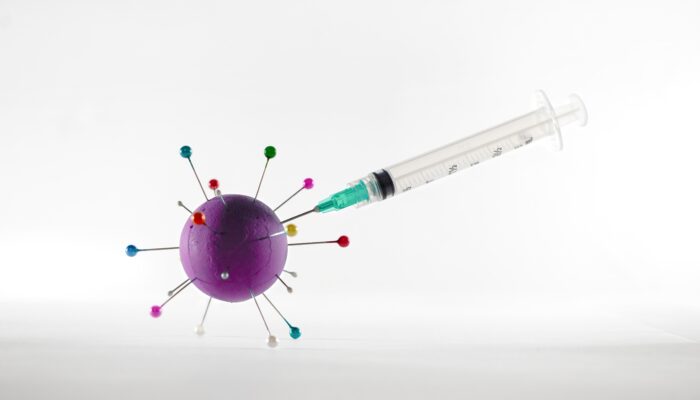 一张照片显示，一个空注射器插在一个冠状病毒模型中，这个模型是用涂成紫色的聚苯乙烯泡沫球制成的，上面有彩色绗缝针，背景是白色。