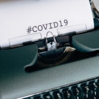 这是一个旧奥林匹亚手动打字机的特写，蓝灰色。里面有一张白纸，上面用大写字母写着COVID - 19。