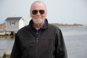 戴着墨镜的微笑男子，白头发白胡子，身穿黑色夹克，站在水边，背景是一座白色的小楼。