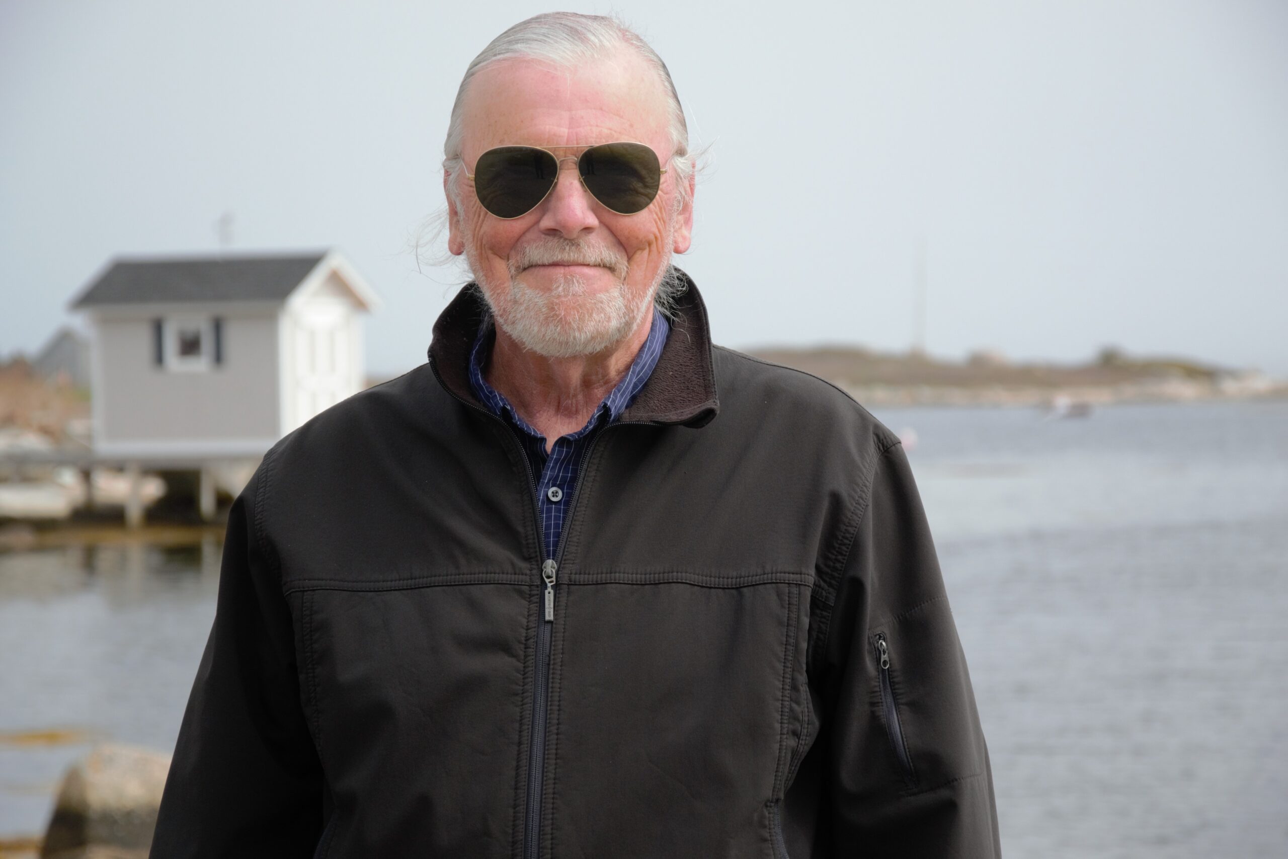 戴着墨镜的微笑男子，白头发白胡子，身穿黑色夹克，站在水边，背景是一座白色的小楼。