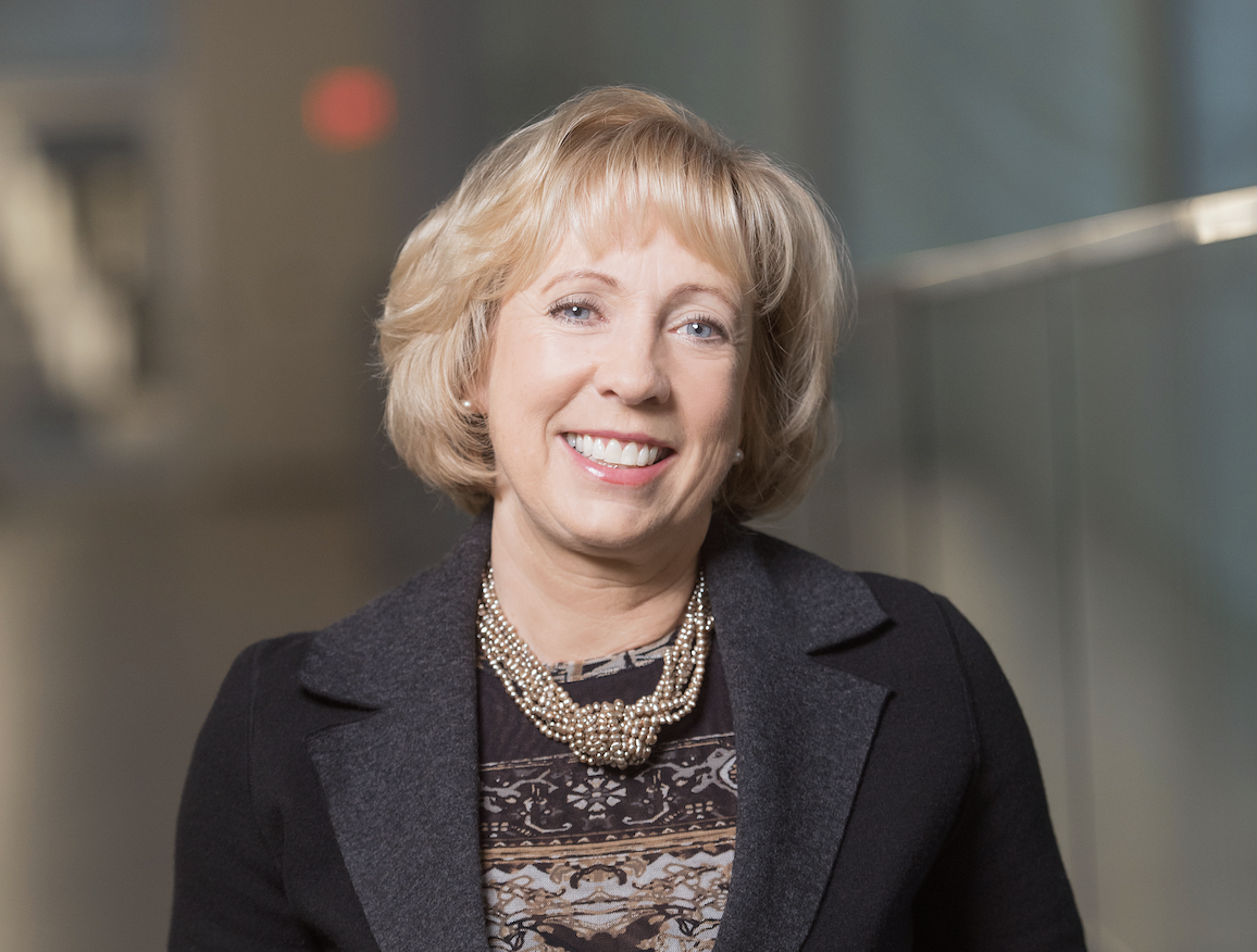 一位留着金色短发、在深棕色夹克里穿着米色和棕色图案的毛衣、面带微笑的女性似乎在礼堂里对着镜头微笑。