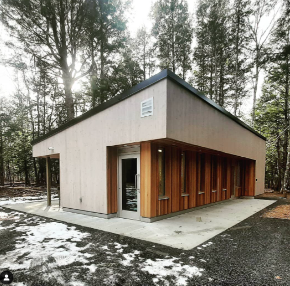 地面上的盥洗室的照片或照片插图，设置在砾石地区的树木之间。它看起来很像弗兰克·劳埃德·赖特(Frank Lloyd Wright)，有粉刷过的木板和天然雪松，还有倾斜的屋顶。