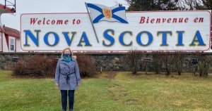 在新不伦瑞克省和新斯科舍省边境，一名下院议员站在欢迎来到新斯科舍省的标志前