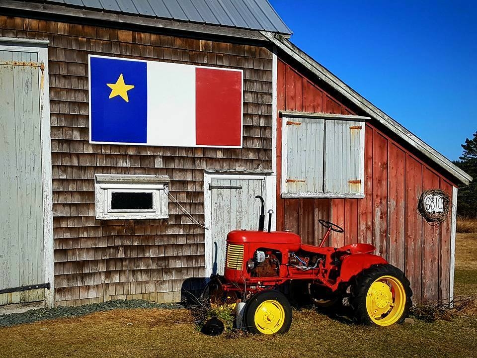 这是一张饱经风霜的谷仓照片，谷仓上挂着鲜艳的阿卡迪亚国旗。牲口棚的一部分是棕色的，有白色的门和装饰，牲口棚还有一处粉刷成红色的附加部分。阿卡迪亚国旗上有蓝色、白色和红色的垂直条纹，蓝色条纹的上半部分有一颗五角星。谷仓前面是一辆鲜红色的老式拖拉机，车轮是鲜黄色的，轮胎是黑色的。