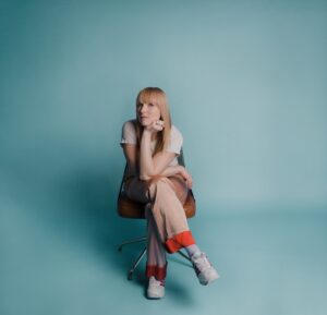 赤港伊莎贝尔照片。她坐在椅子上，用下巴在她的肘部上支撑，她的肘部划船在她的交叉腿上。她穿着时髦的金色米色缎面裤子用一把鲜艳的红色袖口，她的头发是一种类似的颜色，落后的滴水和下方是一个可爱的苍白绿松石。