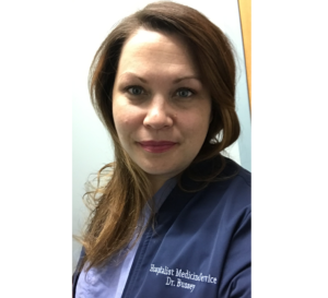 一张Christy Bussy医生的照片。她留着长长的黑发，目不转睛地看着镜头。这张照片是在医院的实验室拍摄的，她穿着一件蓝色或紫色的实验服，胸前写着她的名字和医院名称。