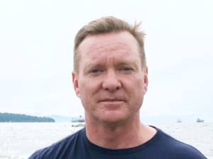 爱德华博士博物馆照片。这是一个明亮而朦胧的夏日。他正直接看着镜头，穿着海军蓝色T恤，有一个严肃的表情。在背景中是像温哥华港一样看起来像温哥华的港口，在远处有几艘集装箱船和渔船。