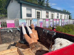 照片上的一个小汽车旅馆建筑与围栏在农村地区。栅栏上有许多旅游海报。前景是一辆小卡车后面的一个涂着红色的大盒子。卡车床上有一袋废弃的白色蕾丝织物，可能是桌布或婚纱。卡车床上还有一个几乎全裸的男性人体模型。他的躯干和下半身是分开的，下半身穿着长长的白色支撑膝袜。他赤裸的假人屁股正对着明亮的阳光。