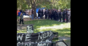 来自视频的屏幕截图，在公园内显示了一个草地。在背景中有一个明亮的蓝色帐篷，以及一大群警察在深色制服，站在路上。一些抗议者是可见的，一个人正在手机上拍摄警察。在前景中是一只手绘牌在公园长凳上，你可以看到我们需要的话，但其余的是模糊的。