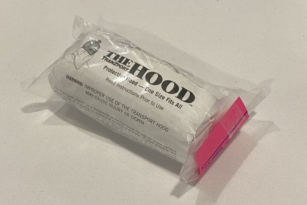 在白色的桌子上看到含有唾液罩的塑料袋。有一个黑白的图，显示一个戴着遮盖嘴和脖子的人的人。引擎盖上的徽标说“The Tranzport引擎盖”。在那些词下，它读了“保护罩 - 一个尺寸适合所有”，然后“，”在使用前阅读说明“。在此之后，它说，在所有帽子上警告：使用的运输不当可能导致伤害或死亡。“右侧包装中包含粉红色的纸张。