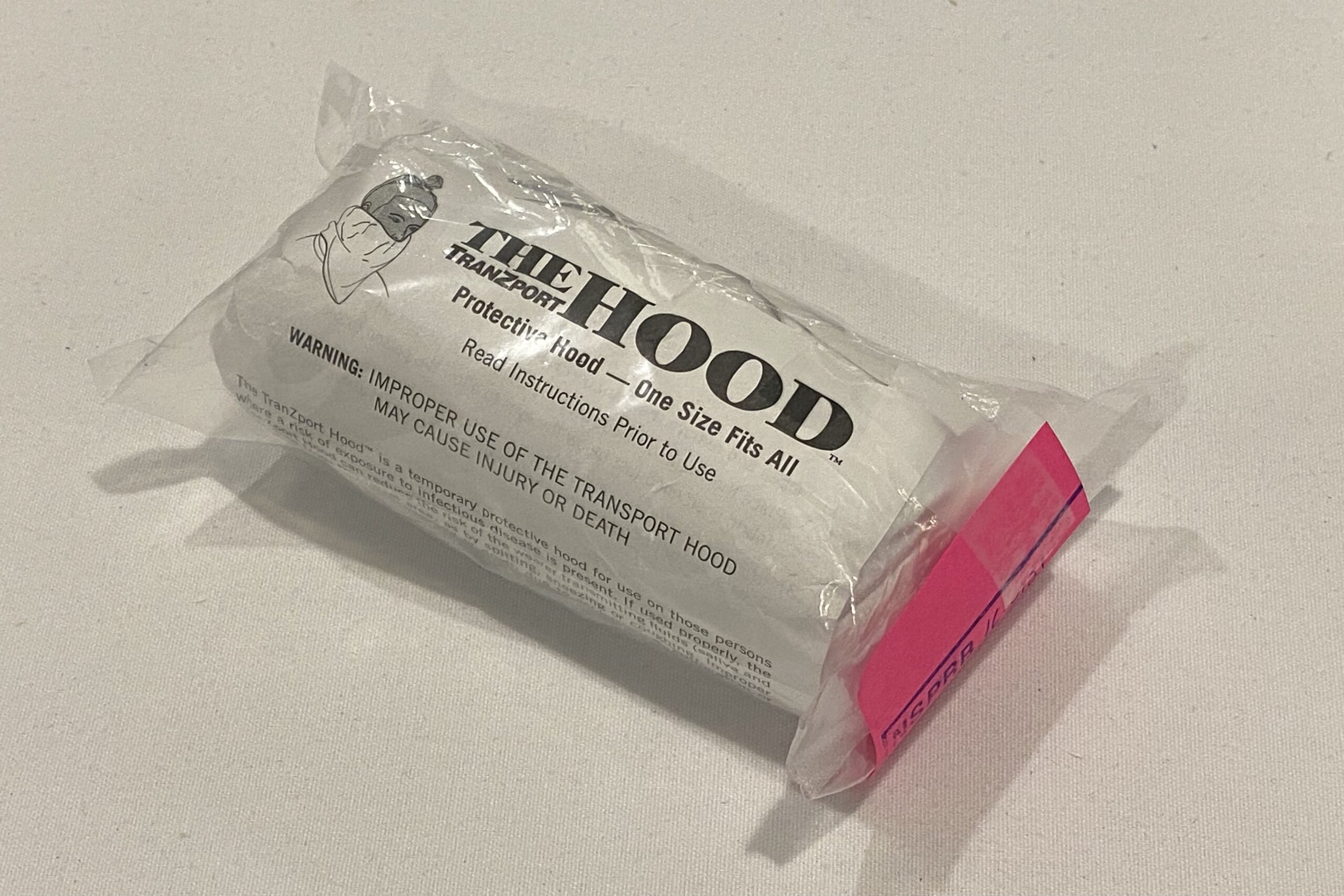 在白色的桌子上看到含有唾液罩的塑料袋。有一个黑白的图，显示一个戴着遮盖嘴和脖子的人的人。引擎盖上的徽标说“The Tranzport引擎盖”。在那些词下，它读了“保护罩 - 一个尺寸适合所有”，然后“，”在使用前阅读说明“。在此之后，它说，在所有帽子上警告：使用的运输不当可能导致伤害或死亡。“右侧包装中包含粉红色的纸张。