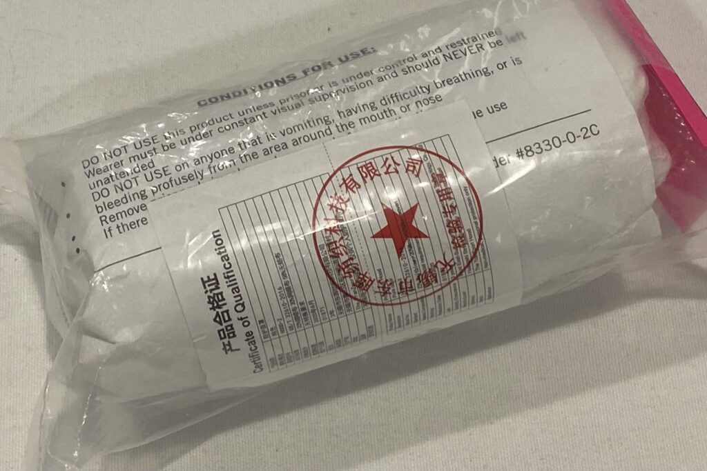 塑料袋的背面在白色的桌子上看到了唾液罩。在包装上有红色的中文写作和一个红星。内部，读取：“使用条件：除非囚犯在控制和抑制中，否则不要使用本产品。佩戴者必须在不断的视觉监督下，不应该无人看管。不要在任何呕吐的人身上使用呼吸困难，或者从口腔或鼻子周围的区域进行流血。“通过包装切断了另外两个指令。