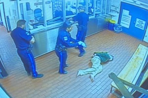 一段模糊的监控视频截图显示，三名身穿制服的警察和一名男子躺在棕色的砖地上。右边是一张长凳，犯人在被登记时通常会坐在那里。在左边的办公室里，警察和售票员之间有一道有机玻璃屏障。