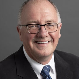 莱尔德·布朗利的照片。他是一个秃顶的男人，灰白的头发，黑框眼镜，穿着深色西装，白衬衫，蓝白条纹领带。他笑得就像有人提醒他今年能挣多少钱一样。