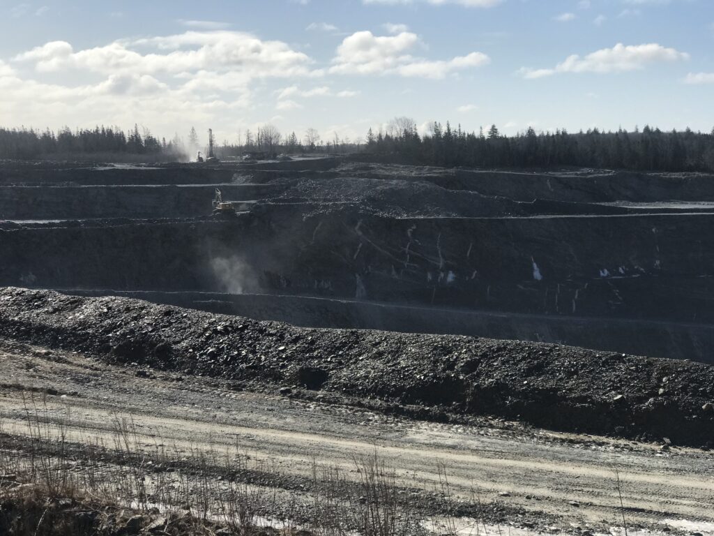 Touquoy露天坑金矿的照片。在大约一公里的地平线上，您可以看到常绿和硬木树的aline剪影，呈现出明亮的蓝天与几个浮肿的云彩。更近的是，您可以看到一些狂欢的Duxt云的设备。在照片的中间是地上令人敬畏的深坑。在前景中是一个带轮胎轨道的干砾石道，地球的肠道堆积在道路和深层坑之间的银行里。