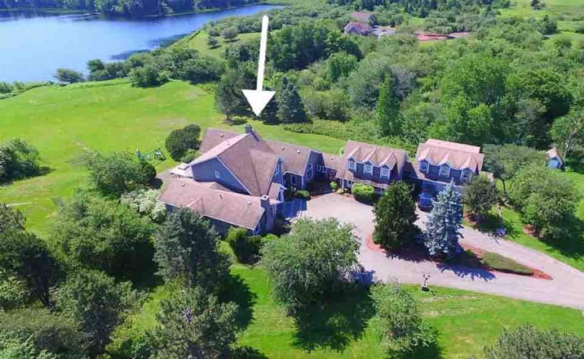 丽塔·麦克尼尔（Rita MacNeil）的故居航拍照片，这是一座可爱的山墙式L形建筑，涂成灰色，白色镶边，坐落在湖边或水湾旁的一处可爱的树木房地产上。