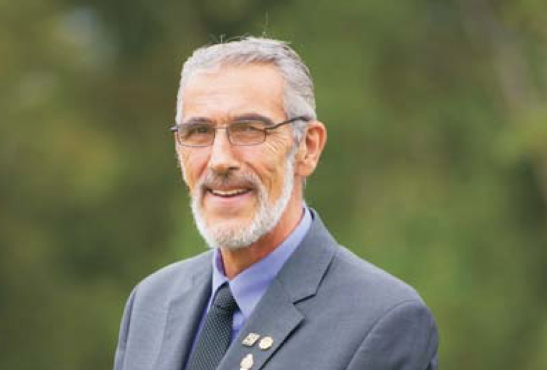 弗农·皮特的一张照片，摄于一个阴天的夏日。有一片模糊的绿树背景。他对着镜头露出狡黠的、斜视的微笑。他穿着灰色西装，淡紫色衬衫和灰色领带。他有三个翻领别针。他头发花白，留着灰色的山羊胡，戴着一副金属框眼镜。