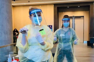 劳伦·米尔斯和她的同事在测试诊所穿着全套个人防护装备的照片。劳伦手里拿着棉签，她的朋友竖起了两个大拇指，他们看起来很高兴能帮忙。