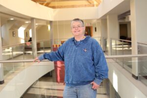 这是Vett Lloyd的照片，摄于建筑内部，有开放式楼梯，来自中庭的光线充满了整个空间。她穿着蓝色运动衫和牛仔裤，对着镜头微笑。