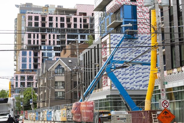 2021年6月，哈利法克斯克莱德街在建的公寓楼。新建筑使一座只有几十年历史的老建筑相形见绌。在前景中，你可以看到一个亮蓝色的樱桃采摘起重机，和一个黄色的管道，垃圾通过它落入垃圾箱。在最近的建筑前面是一个铁链围栏，上面覆盖着各种颜色的横幅，颂扬建筑公司、投资者和未来的豪华住宿。横幅已经破旧不堪了。