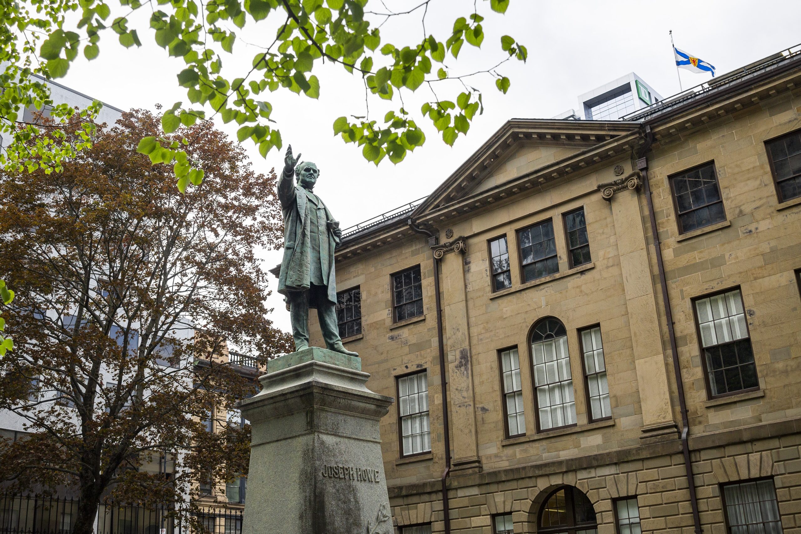 一座历史悠久的石头建筑前，一座男子雕像在基座上挥手。雕像靠近一棵枝繁叶茂的树