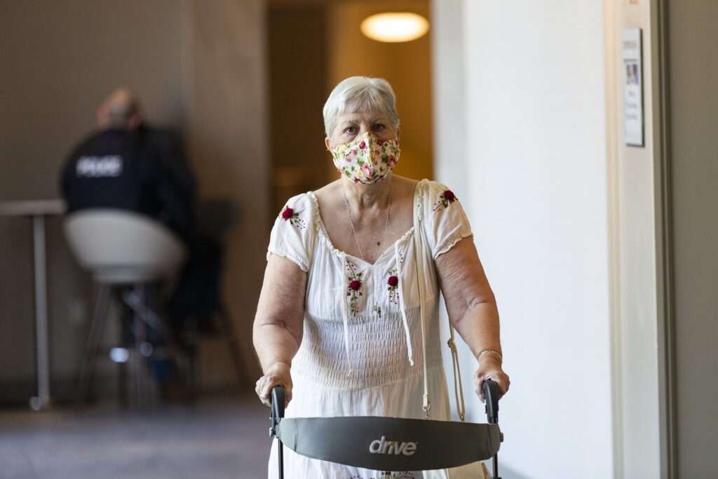一个女人用助行器走过酒店的走廊，去参加警察审查委员会的听证会。她穿着一件白色的连衣裙，戴着一个花布面具。