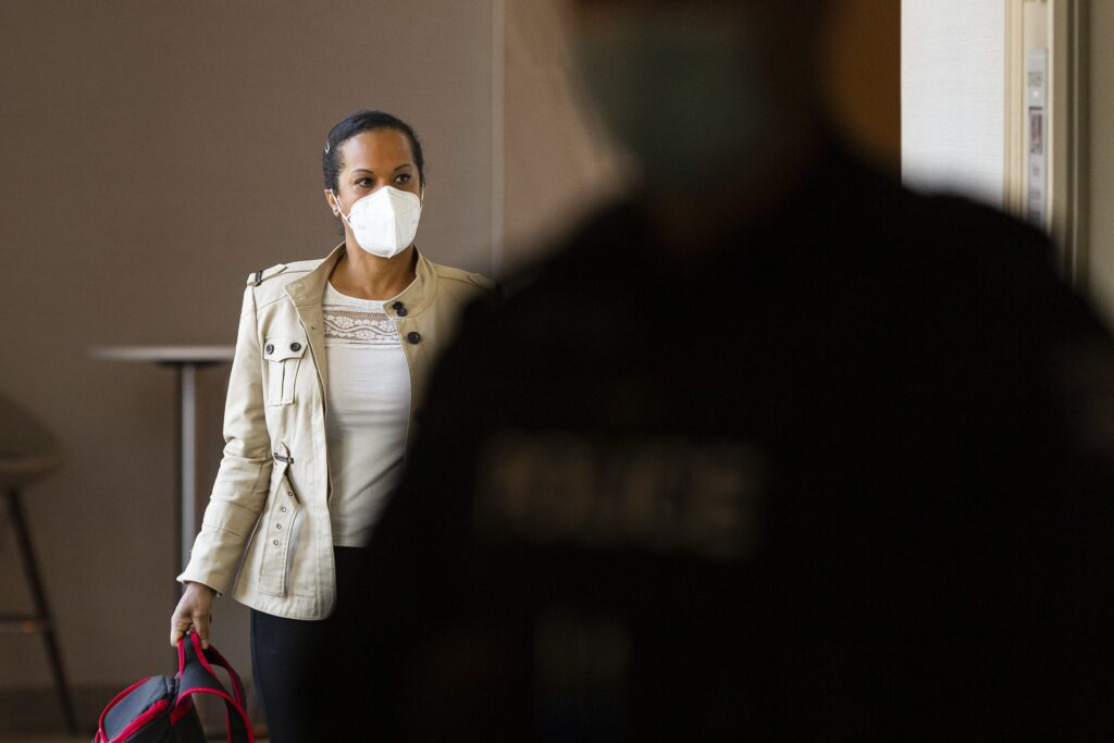 一名戴着白色面具、穿着棕色夹克、背着背包的女子朝左边看，走在酒店的走廊上。在前景中，一个戴着面具的模糊警察穿过现场。